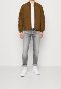 Brun, kviltad bomberjacka med ribbad krage, kombinerad med ljusgrå slim-fit jeans och vita sneakers. En enkel vit t-shirt under.