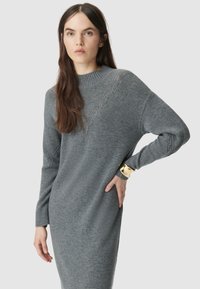 Szara dzianinowa sukienka z wysokim kołnierzem, długimi rękawami i prążkowanymi mankietami. Posiada przekątny wzór tekstury i luźny krój. Złota bransoletka na mankietach.