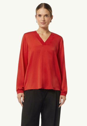 Rote Bluse mit langen Ärmeln aus glattem, leichtem Stoff, mit V-Ausschnitt und gerafften Manschetten, kombiniert mit schwarzen, locker sitzenden Hosen.