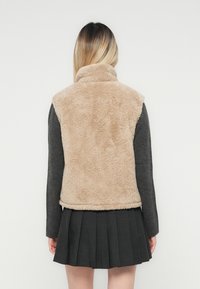 Vrouw in een beige mouwloze fleece vest over een donkere lange mouw top en een donkere geplooide rok, gezien van achteren tegen een witte achtergrond.
