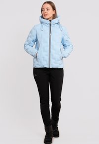 Helles blaues Pufferjacke mit hohem Kragen, Frontreißverschluss und Kordelzügen. Verfügt über Seitentaschen und eine gesteppten Struktur. Getragen mit schwarzen Hosen und Stiefeln.