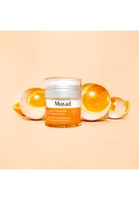 Murad Essential-C Overnight Barrier Repair Cream i en klar burk med orange accenter; omgiven av glasapelsiner på en persikofärgad bakgrund.