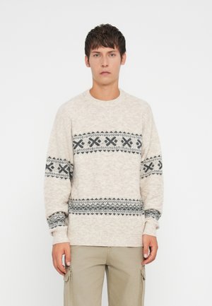 Junger Mann in einem beigen Pullover mit grauen geometrischen Mustern und beigen Cargo-Hosen, stehend vor einem schlichten weißen Hintergrund.