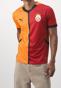 Camiseta de fútbol a rayas rojas y naranjas con acentos en negro, con un cuello en V, logo de Puma y emblema del club con cuatro estrellas.
