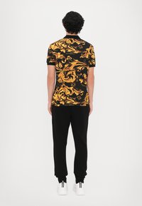 Camisa de manga curta com padrão preto e dourado, com desenhos em espiral. Combinada com calças pretas e ténis brancos. Vista traseira, tecido texturizado.