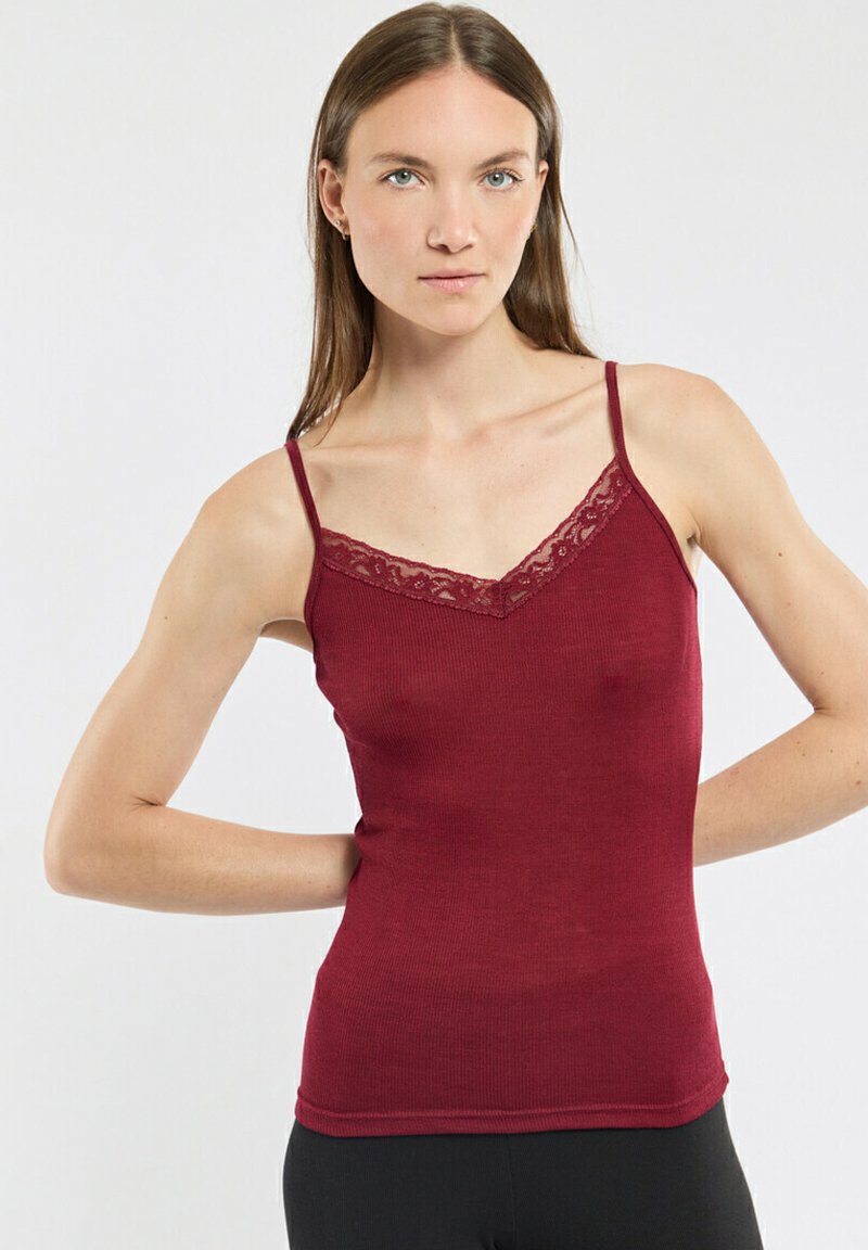 Femme aux cheveux bruns raides portant un top sans manches bordeaux avec des bordures en dentelle et un pantalon noir, se tenant les mains derrière le dos.