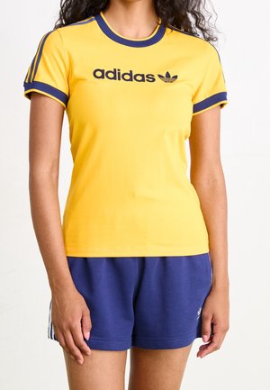 Gul bomulds T-shirt med marineblå detaljer, rund halsudskæring og korte ærmer. Har et sort "adidas" logo og ikonisk trekløverdesign.