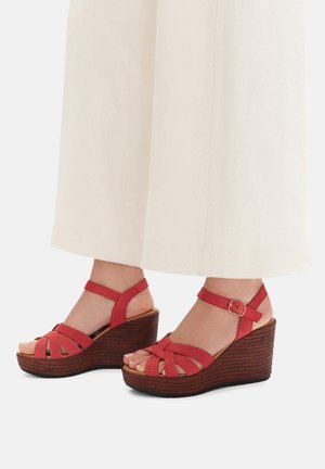 Sandalias cuña de cuero rojo con diseño trenzado, tiras ajustables en el tobillo y textura de madera en el tacón de cuña.