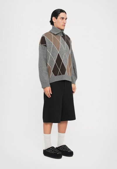 Sudadera gris con un patrón de diamantes en marrón y beige, combinada con pantalones cortos negros y zapatillas bordadas negras. Modelo de pie.