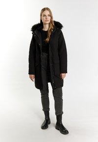 Sort parka-frakke med faux fur hætte, lynlås og knaplukninger, to forlommer, parret med grå jeans og sorte ankelstøvler.