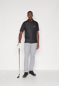 Svart vadderad väst över en kortärmad svart tröja, ljusgrå byxor, svarta golfskor och håller en golfklubba. Gul logotyp på västen.