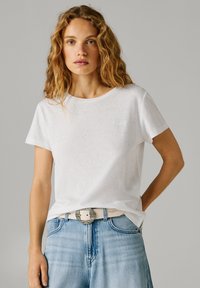 Kvinde med bølget blondt hår iført en simpel hvid t-shirt, stoppet ned i lyseblå jeans med et hvidt bælte med en dekorativ sølvspænde.