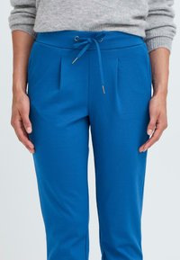 Blaue Hose mit glatter Textur, gefaltetem Vorderteil und Kordelzugbund. Merkmale sind metallische Akzente am Kordelzug und eine taillierte Silhouette.