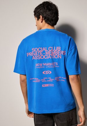 Modrá bavlnená tričko s veľkým ružovým textom na chrbte, ktorý zahŕňa "SOCIAL CLUB PRIVATE-MEMBERS ASSOCIATION" a rôzne názvy miest.