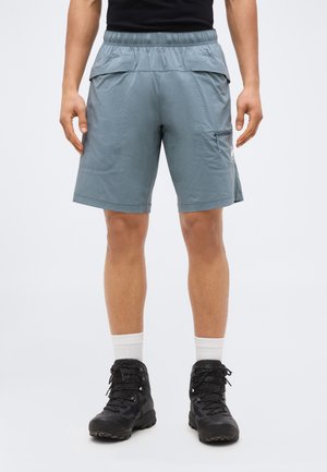 Άνδρας που φοράει ανοιχτό μπλε σορτς πεζοπορίας με πίσω τσέπες με καπάκι, πλευρική τσέπη με φερμουάρ, λευκές κάλτσες και μαύρα μποτάκια πεζοπορίας, στέκεται μπροστά σε απλό φόντο.