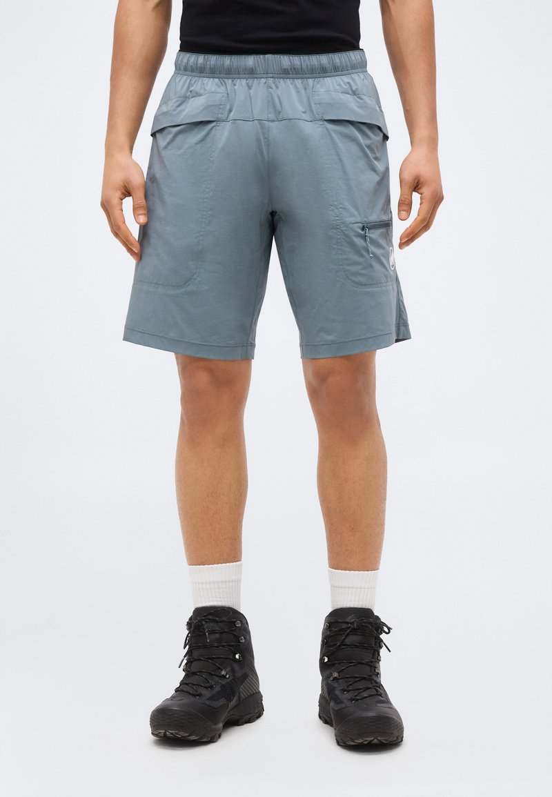 Hombre vistiendo pantalones cortos de senderismo azul claro con bolsillos traseros con solapas, bolsillo lateral con cremallera, calcetines blancos y botas de senderismo negras, de pie contra un fondo liso.