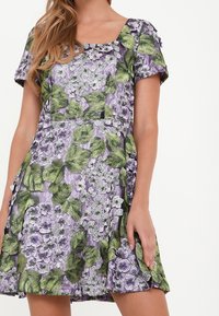 Blommig klänning med fyrkantig halsringning, korta ärmar och lila tyg med mönster av gröna blad och vita blommor.