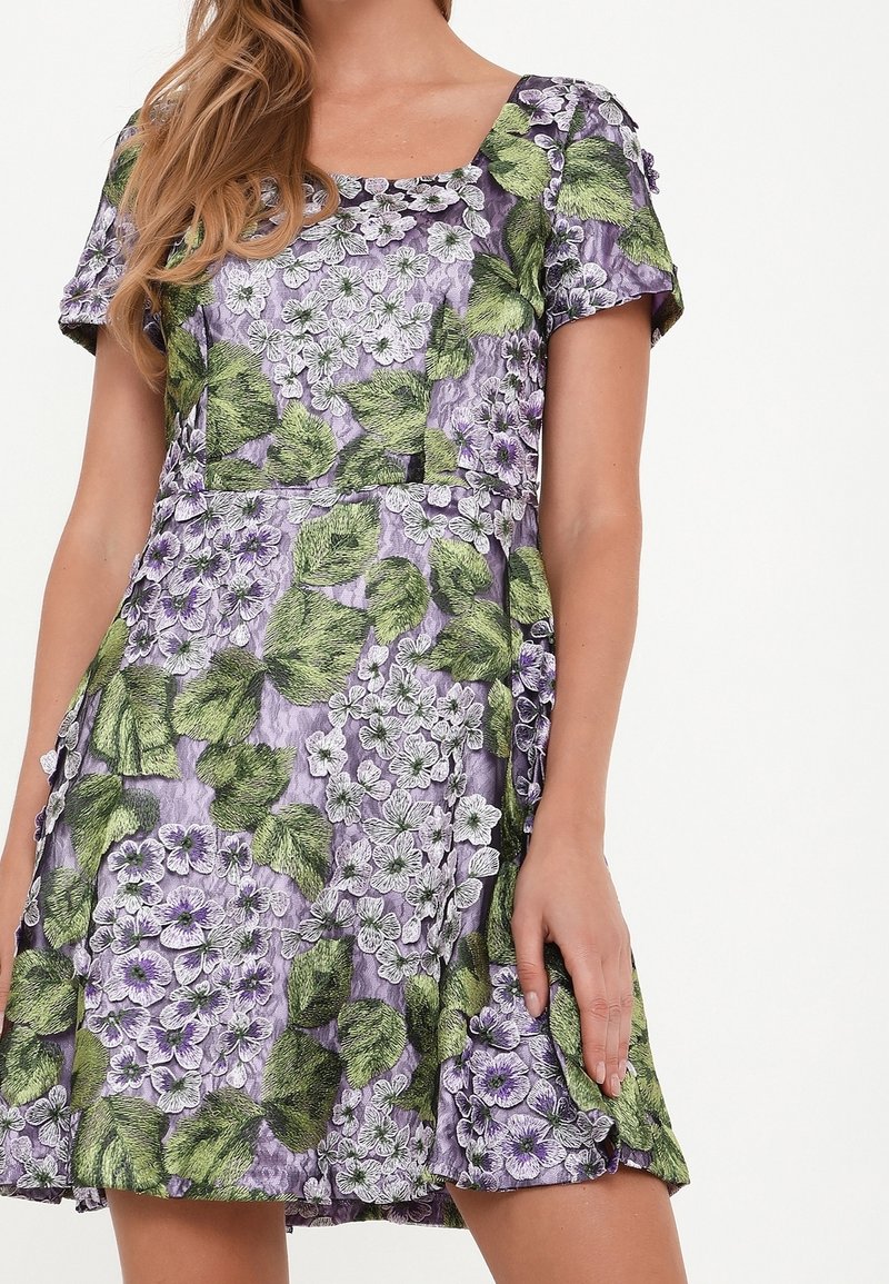Blommig klänning med fyrkantig halsringning, korta ärmar och lila tyg med mönster av gröna blad och vita blommor.