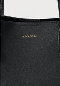 Čierna kabelka ztextúrovaného syntetického materiálu s predným zlatým logom „ANNA FIELD“ a dvojitými rukoväťami.
