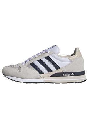 Baskets Adidas blanches avec trois bandes noires, détails en suède beige, lacets blancs, semelle noire et logo Adidas au talon.