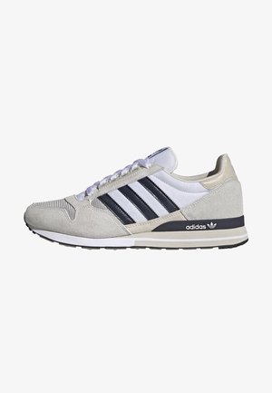 Witte Adidas-sneaker met drie zwarte strepen, beige suèdedetails, witte veters, zwarte zool en Adidas-logo op de hiel.