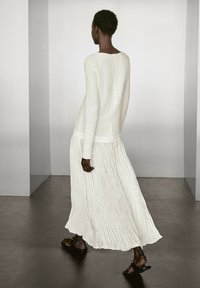 Langes weißes Kleid aus strukturiertem Stoff mit einem ausgestellten Rock, kombiniert mit schlichten schwarzen Sandalen. Das Design hat einen bescheidenen Ausschnitt und lange Ärmel.