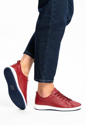 Person wearing dark blue jeans and red leather sneakers with white soles, one foot raised showing navy blue tread on sole.