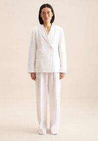 Witte linnen dubbelgeknopte colbert met twee voorkeurszakken, gecombineerd met losse witte linnen broek met een gestructureerde afwerking.
