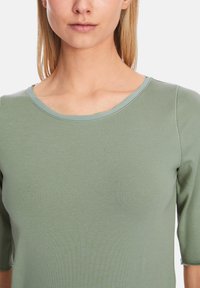 Kurzärmeliges Oberteil aus lichtgrünem Stoff, mit rundem Ausschnitt und sichtbaren Nähten entlang der Kanten. Glatte Textur.