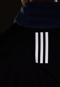 Svart sportsjakke med høy krage og tre vertikale hvite striper på øvre del av ryggen. Materialet ser glatt og lett ut.