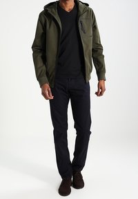 Muž v olivově zelené kapuci, černém tričku s výstřihem do V, tmavě modrých kalhotách a tmavohnědých botách stojící na bílém pozadí.