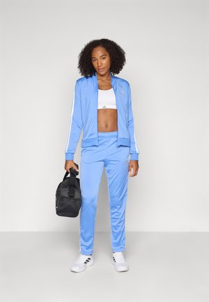 Kohutunud juustega naine, kes kannab helesinist dressipluusi, valget spordirinnahoidjat ja valgeid tossusid, hoides musta jõusaalikotti, seisab ühevärvilise heleda tausta ees.