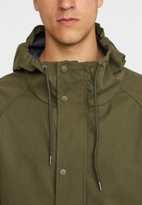 Olivgrüne Kapuzenjacke mit einem Frontreißverschluss, Druckknopfverschlüssen, Kordelzug und kontrastierender Innenauskleidung. Der Stoff wirkt strukturiert und robust.