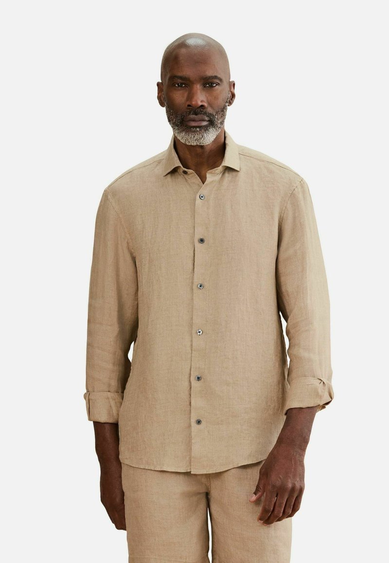 Homme à la barbe grise portant une chemise beige à manches longues boutonnée et un pantalon assorti, debout devant un fond blanc uni.