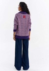 Kobieta zwrócona tyłem, ubrana w sweter w jodełkę w kolorach fioletowym i różowym z naszywką "Miłośnicy książek" oraz szerokie ciemnoniebieskie spodnie.