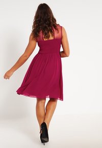 Žena s dlouhými, vlnitými vlasy, oblečená v těsných, burgundských šatech a černých podpatcích, se otáčí, čímž ukazuje volnou sukni šatů.