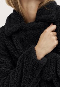 Zwarte, donzige fleece badjas met een brede kraag en omgeslagen manchetten, die een textuurrijk stof laat zien. Hand die de stof van de badjas bij de kraag vasthoudt.