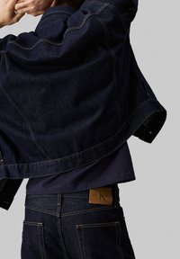 Veste en denim noir au design fluide, dotée de coutures contrastantes. Portée sur un haut sombre, avec un patch en cuir visible sur le jean.