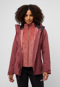 Veste zippée bordeaux avec capuche, portée par-dessus une couche en polaire rouge clair. La veste comporte des poches latérales, des fermetures velcro et une texture lisse.