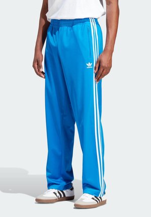 Osoba ubrana w jasnoniebieskie spodnie dresowe Adidas z białymi paskami po bokach, białe trampki z czarnymi paskami i zwykłą białą koszulkę.