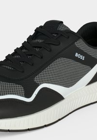 Chaussure de sport en mesh noir et gris avec lacets noirs, semelle blanche et logo « BOSS » sur le côté, vue en gros plan.