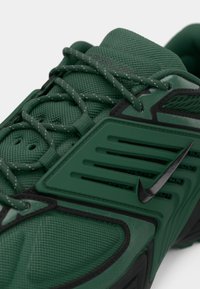 Gros plan sur une chaussure de sport verte avec un tissu texturé, des accents noirs, des lacets rayés et un swoosh Nike noir sur le panneau latéral.
