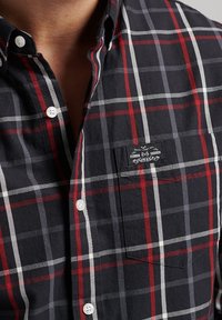 Camicia a quadri in nero, rosso e bianco; tessuto di cotone; colletto con bottoni; taschino sul petto sinistro con logo; bottoni bianchi; motivo a griglia distintivo.