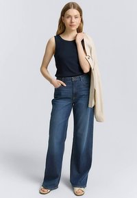 Haut sans manches bleu marine associé à un jean taille haute à jambes larges bleu foncé. La tenue est complétée par une veste beige clair. Textures lisses.