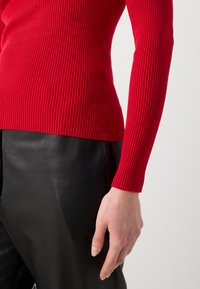 Haut à manches longues rouge à côtes, fabriqué dans un matériau extensible, associé à un pantalon en cuir noir, présentant un gros plan de la manche et de la main.