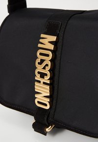 Must must kott musta nailonist, millel on sile tekstuur, küljes kuldne logoaken ja kunstsuede rihm lisadetailina. Kompaktne ristküliku kuju.