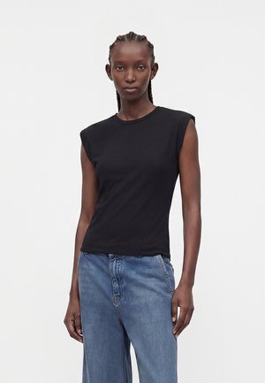 Vrouw met gevlochten haar die een zwart mouwloos topje met gewatteerde schouders en blauwe hooggetailleerde jeans draagt, staande tegen een effen achtergrond.