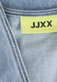 Plava traper tkanina s vidljivim tkanjem, žuta oznaka s natpisom "JJXX" u crnom tekstu i kontrastni narančasti šavovi duž rubova.