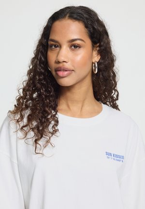 Kvinde med langt krøllet hår iført sølv hoops og en hvid skjorte med blå "SUN KISSED" tekst på brystet.