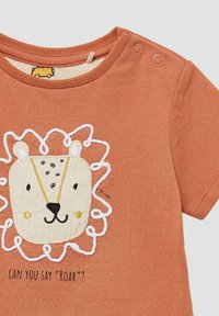 Orange T-Shirt mit kurzen Ärmeln, das ein Löwengesicht-Design mit bestickten Details zeigt, einschließlich einer weißen Mähne und dem Text "KANNST DU 'ROAR' SAGEN?"